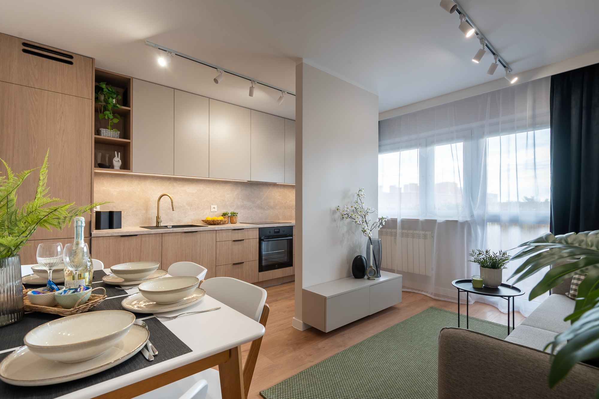 Jasne, przestronne wnętrze: kuchnia z drewnianymi szafkami i marmurowym blatem, jadalnia z nakrytym stołem i salon z zielonym dywanem. Oświetlenie punktowe.