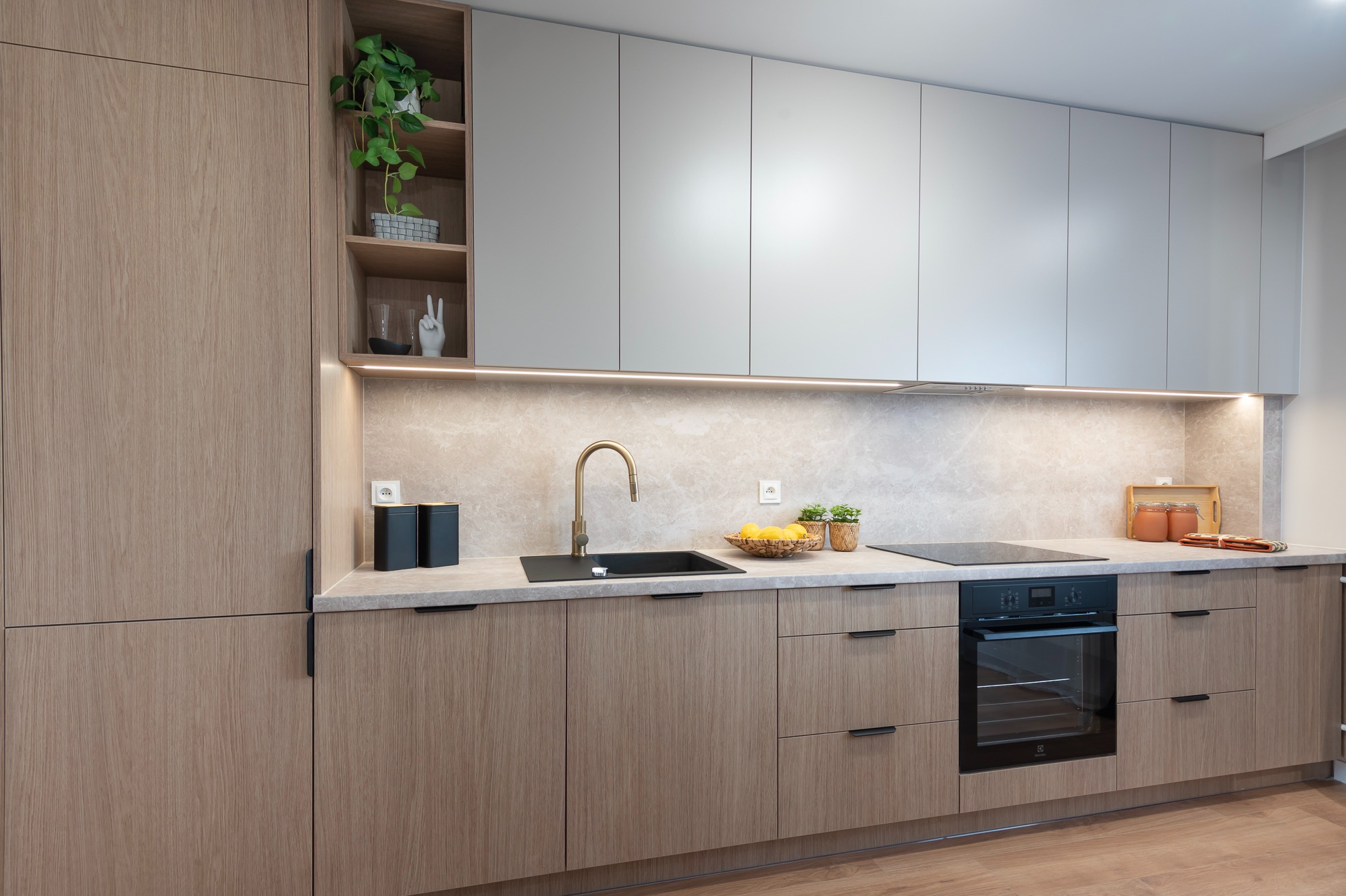 Nowoczesna kuchnia z drewnianymi frontami i szarymi szafkami wiszącymi. Złota bateria, czarny zlew i płyta indukcyjna. Oświetlenie LED pod szafkami.