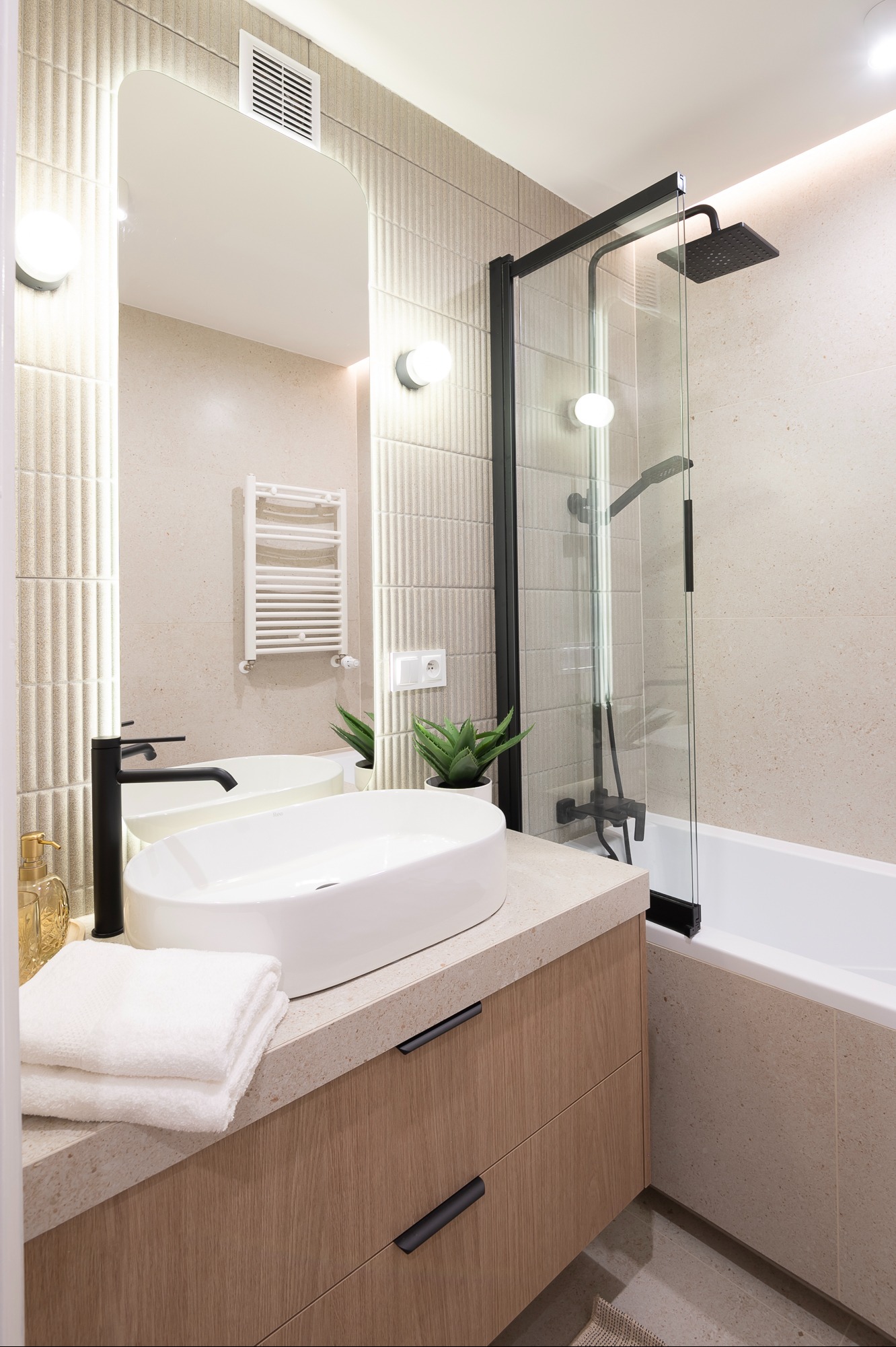 Jasna łazienka z owalną umywalką, czarną armaturą i kabiną prysznicową. Na blacie leżą białe ręczniki, a w lustrze odbija się grzejnik drabinkowy.
