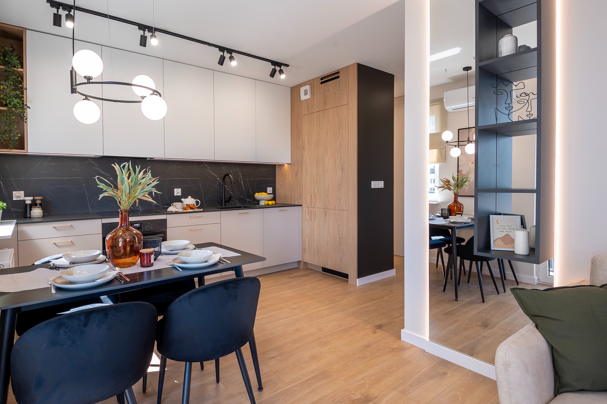 Nowoczesna kuchnia z jadalnią: beżowe szafki, czarny blat marmurkowy, drewniane akcenty, okrągła lampa i stół z czarnymi krzesłami. Lustro powiększa przestrzeń.