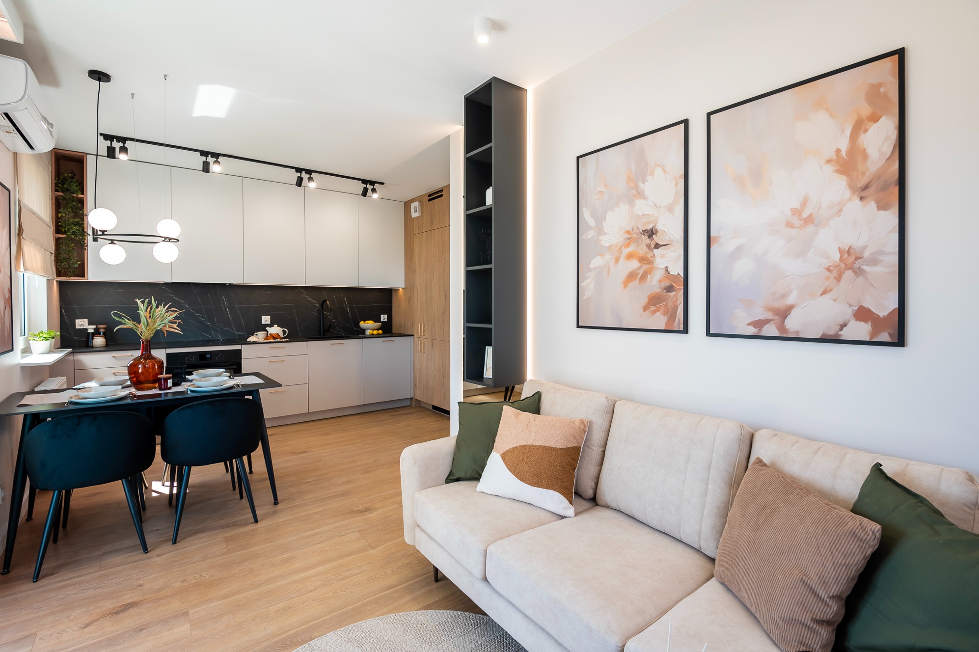 Jasne wnętrze mieszkania po remoncie: salon z sofą i obrazami, jadalnia z czarnym stołem i kuchnia z marmurowym blatem. Nowoczesne oświetlenie punktowe.