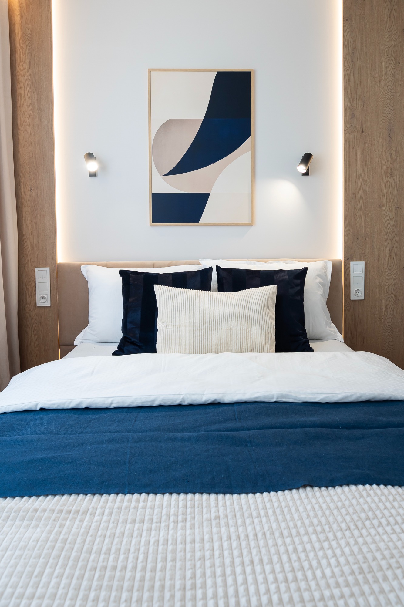 Nowoczesna sypialnia z łóżkiem, obrazem i oświetleniem LED. Drewniane panele ścienne z wbudowanymi lampkami. Minimalistyczny design z akcentami bieli i granatu.