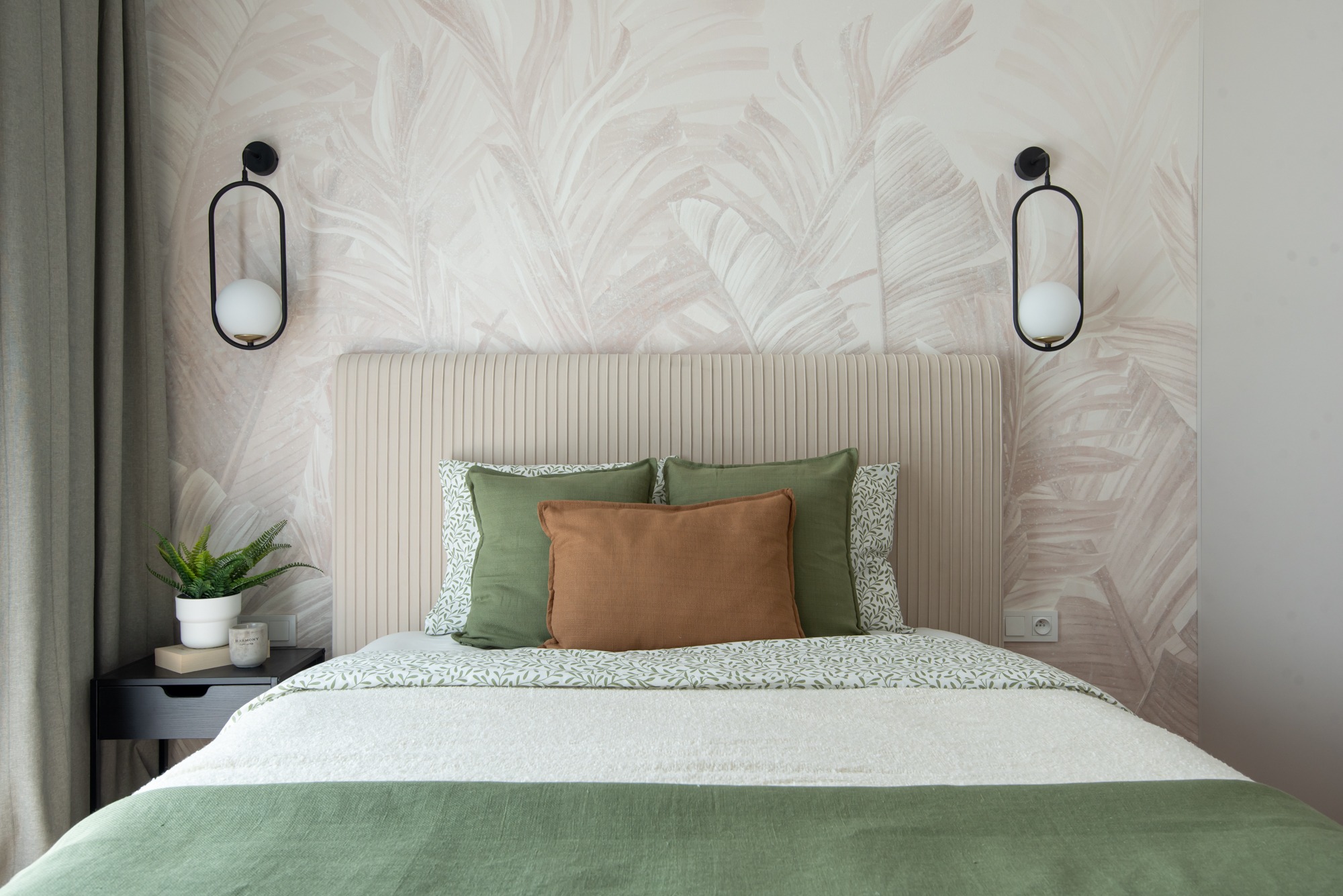 Stylowa sypialnia z łóżkiem, beżowym zagłówkiem i zielonymi akcentami. Na ścianie tapeta w liście i lampy ścienne. Na szafce nocnej roślina w doniczce.