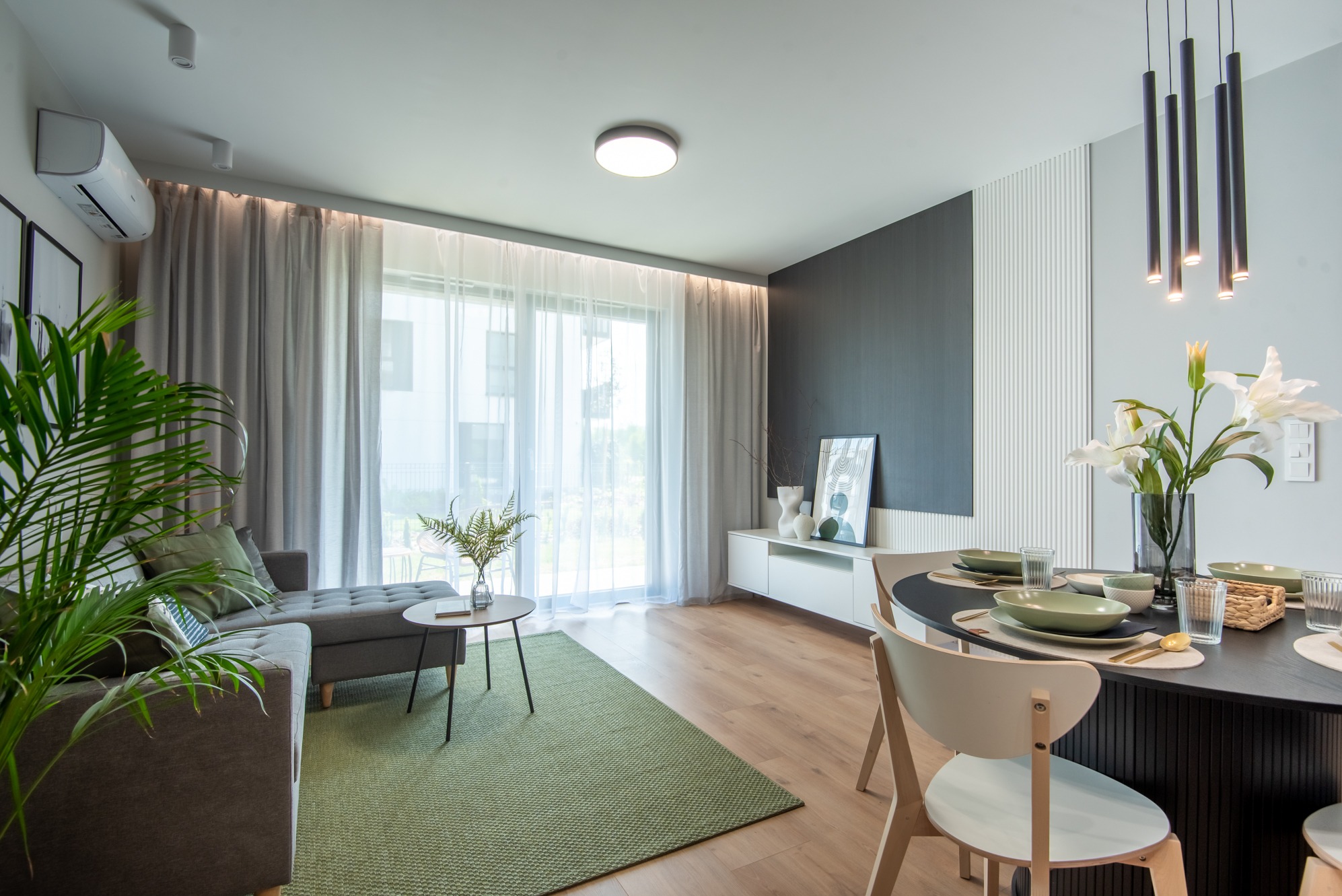 Jasne wnętrze salonu z jadalnią: szary narożnik, zielony dywan, okrągły stół z nakryciem. Minimalistyczny design, nowoczesne oświetlenie, duże okno z zasłonami.