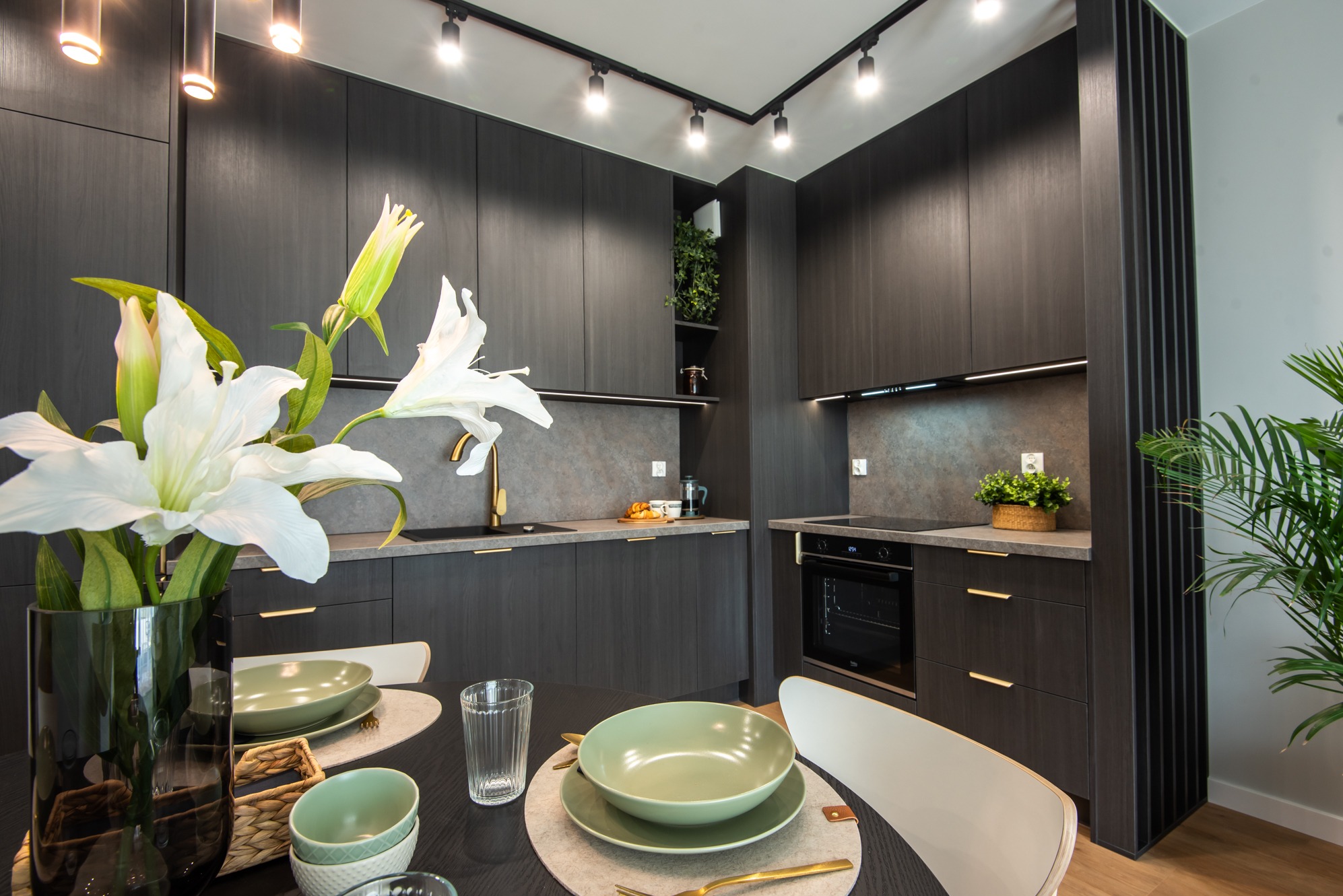 Nowoczesna, ciemna kuchnia z wyspą i kwiatami w wazonie, oświetlenie punktowe, Kraków. Minimalistyczny design, drewniane fronty, złote akcenty.