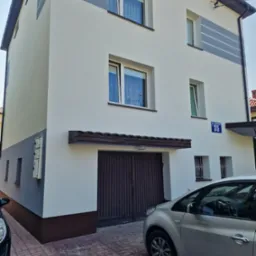 Dwupiętrowy dom po renowacji elewacji: białe ściany z szarymi akcentami, brązowy cokół i brama garażowa. Widoczny fragment chodnika z kostki i zaparkowane samochody.