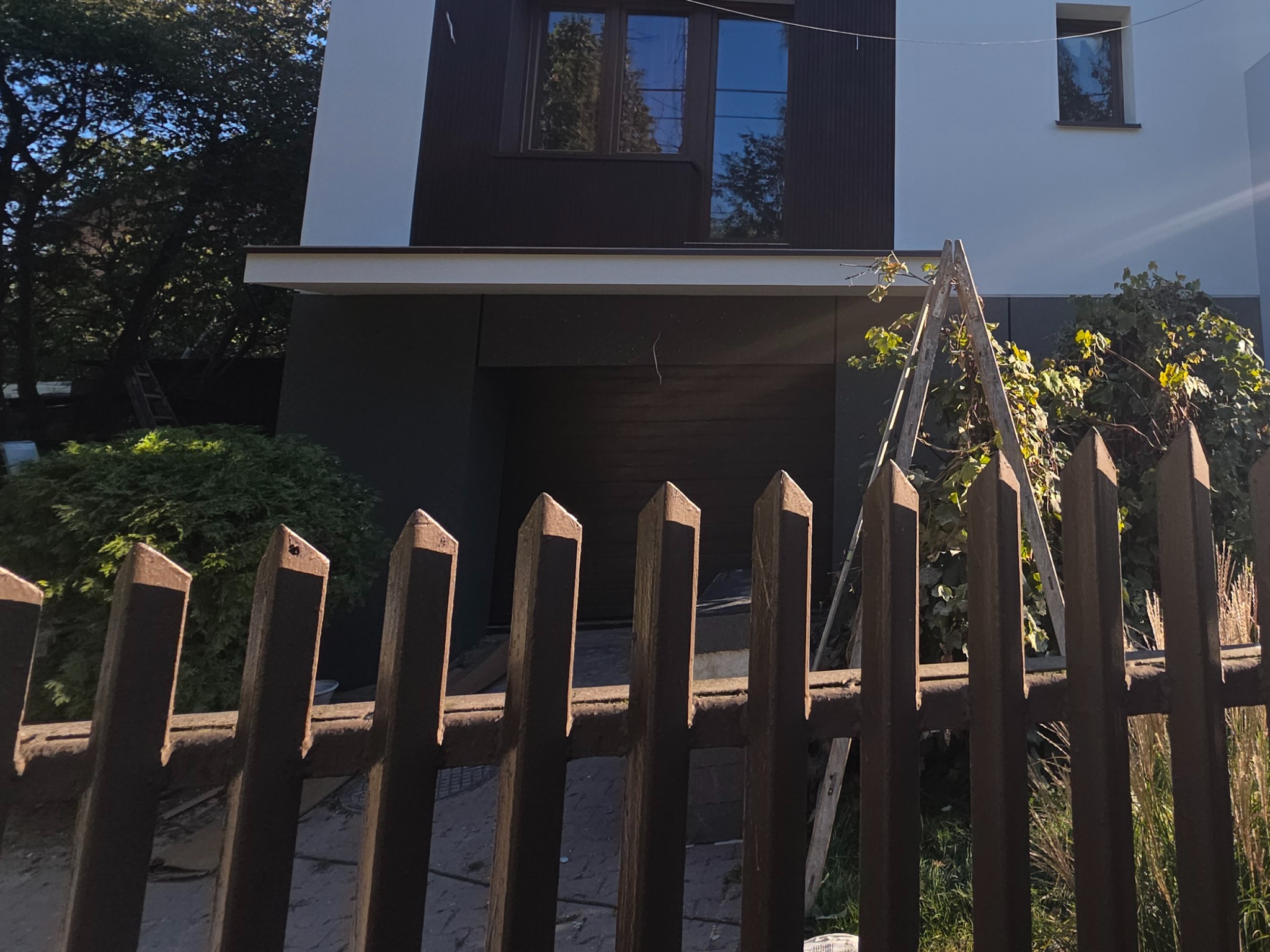 Nowoczesny dom jednorodzinny z ciemną elewacją i białymi akcentami, widziany zza drewnianego płotu. Drabina oparta o ścianę, sugerująca prace wykończeniowe.