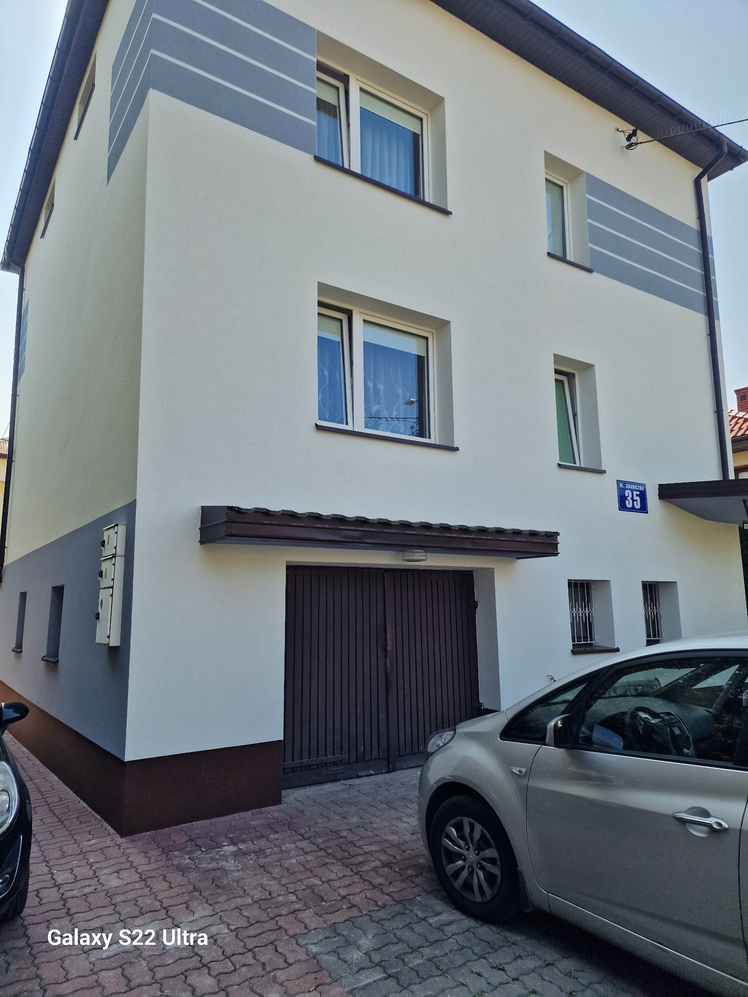 Dwupiętrowy dom po renowacji elewacji: białe ściany z szarymi akcentami, brązowy cokół i brama garażowa. Widoczny fragment chodnika z kostki i zaparkowane samochody.