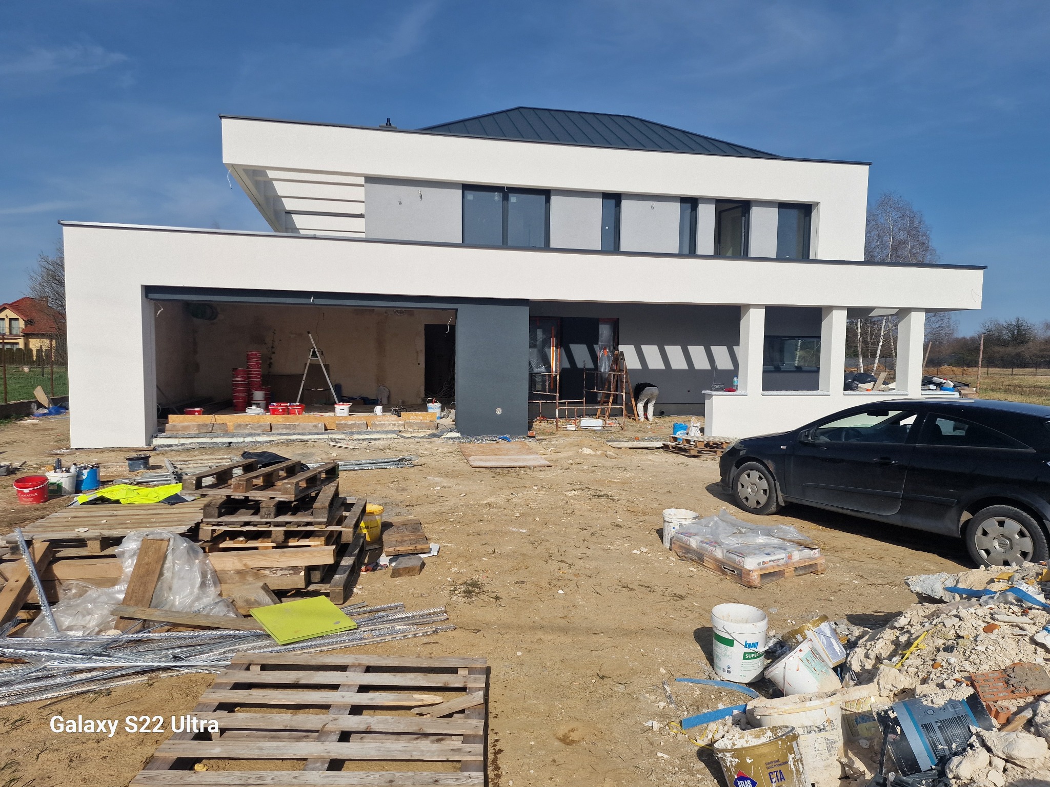 Nowoczesny dom w trakcie prac elewacyjnych: świeżo otynkowana, biała fasada, widoczne rusztowania i materiały budowlane. Czarny samochód na tle budynku.