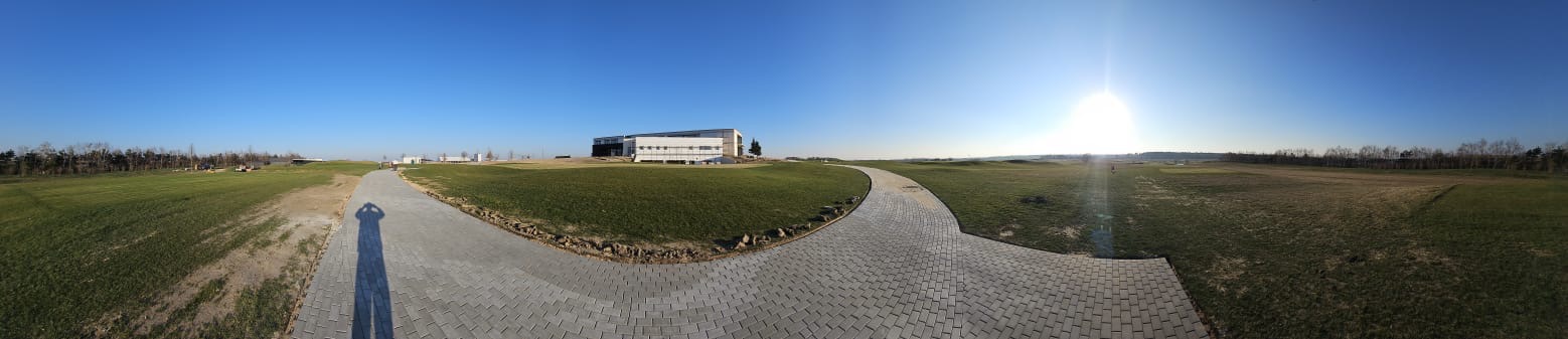 Panoramiczny widok na ścieżkę z kostki brukowej prowadzącą do nowoczesnego budynku na tle zieleni i błękitnego nieba z cieniem osoby robiącej zdjęcie.