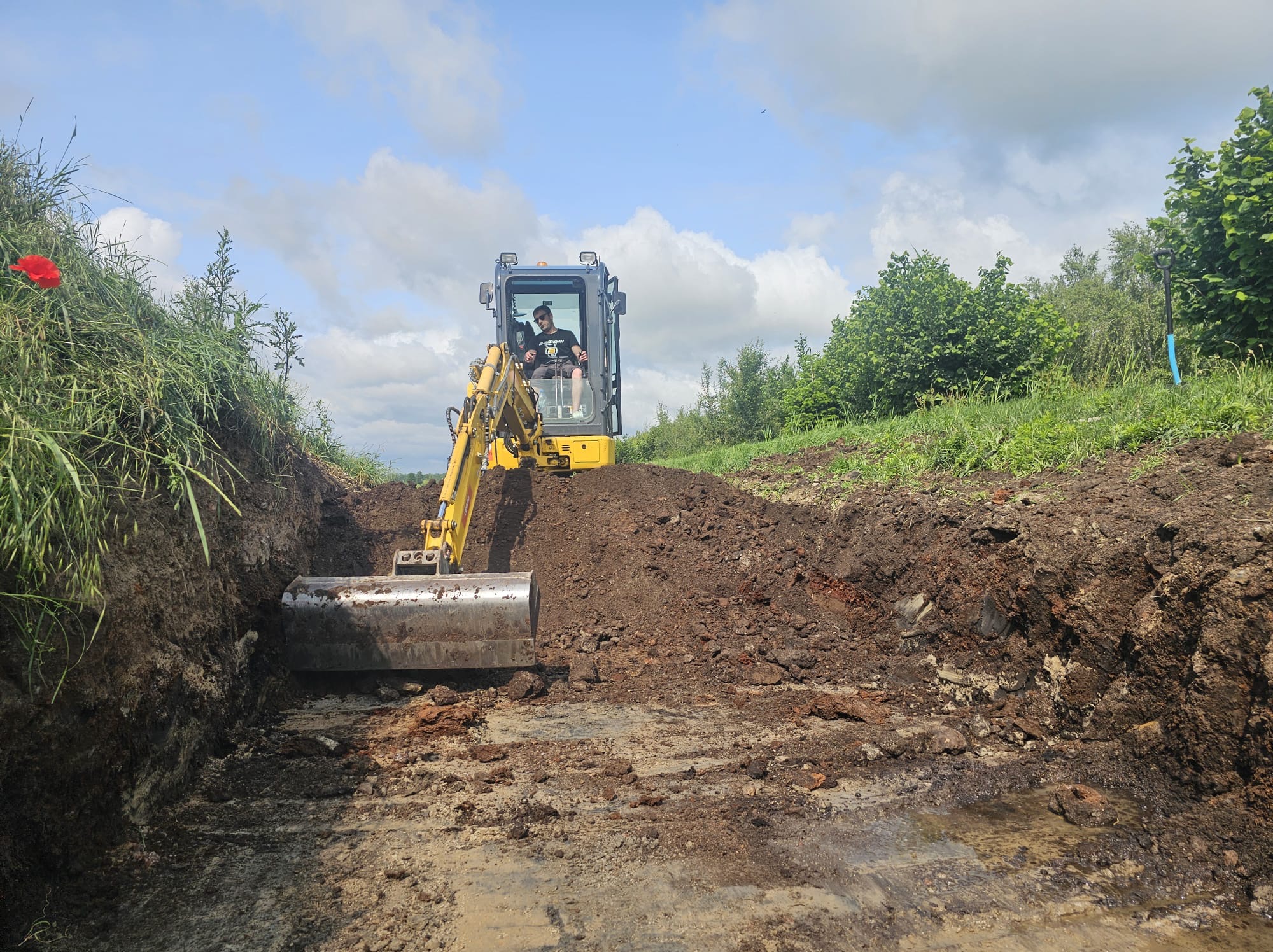 Żółta koparka w trakcie prac ziemnych, operator w kabinie, wykopany rów w ziemi, zielona roślinność w tle, błękitne niebo.