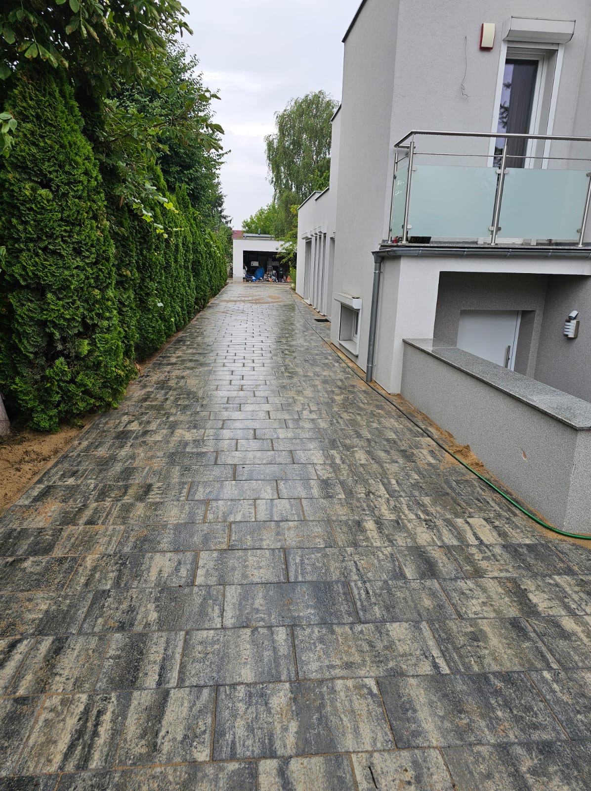 Ułożona kostka brukowa w odcieniach szarości prowadząca do garażu, obok domu z balkonem i żywopłotu. Widok z perspektywy.