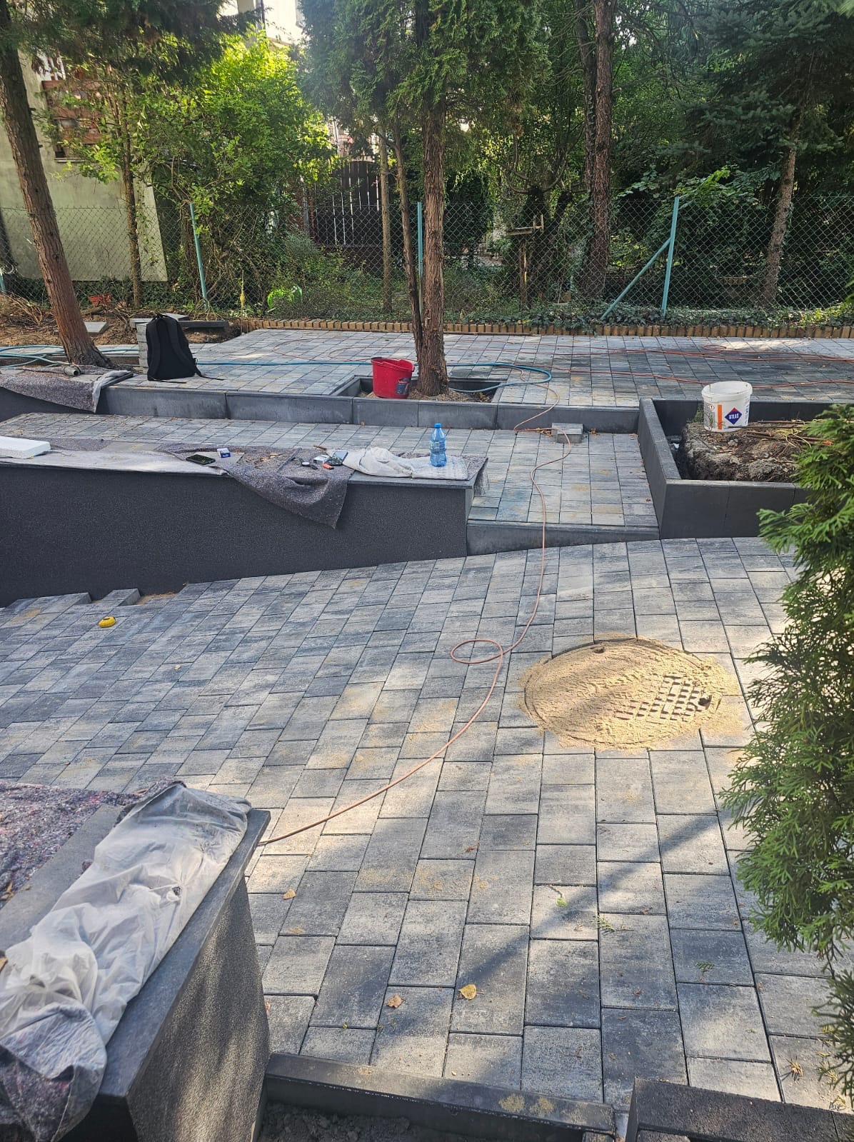 Nowa nawierzchnia z kostki brukowej w ogrodzie, z wykończonymi obrzeżami i miejscami na drzewa. Widoczny właz kanalizacyjny i narzędzia pracy.