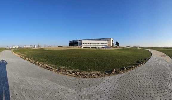 Panoramiczny widok na nowoczesny budynek z kostki brukowej prowadzącej przez trawnik, błękitne niebo w tle. Cień osoby w lewym dolnym rogu.