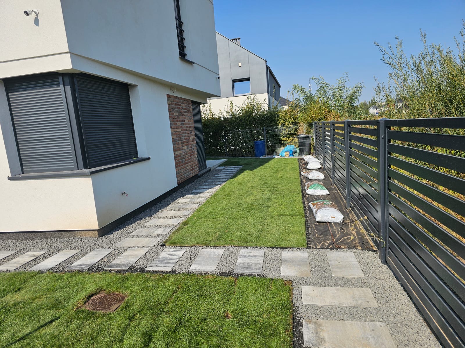 Nowoczesny ogród z trawnikiem w rolkach, ścieżką z płyt i szarego żwiru, obok domu z ceglanym akcentem i czarnym ogrodzeniem panelowym. Worki z ziemią w tle.