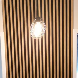 Panel ścienny z pionowych listew w kolorze drewna i czerni, oświetlony nowoczesną lampą wiszącą. Dekoracja wnętrza z wazonem i suszem na pierwszym planie.