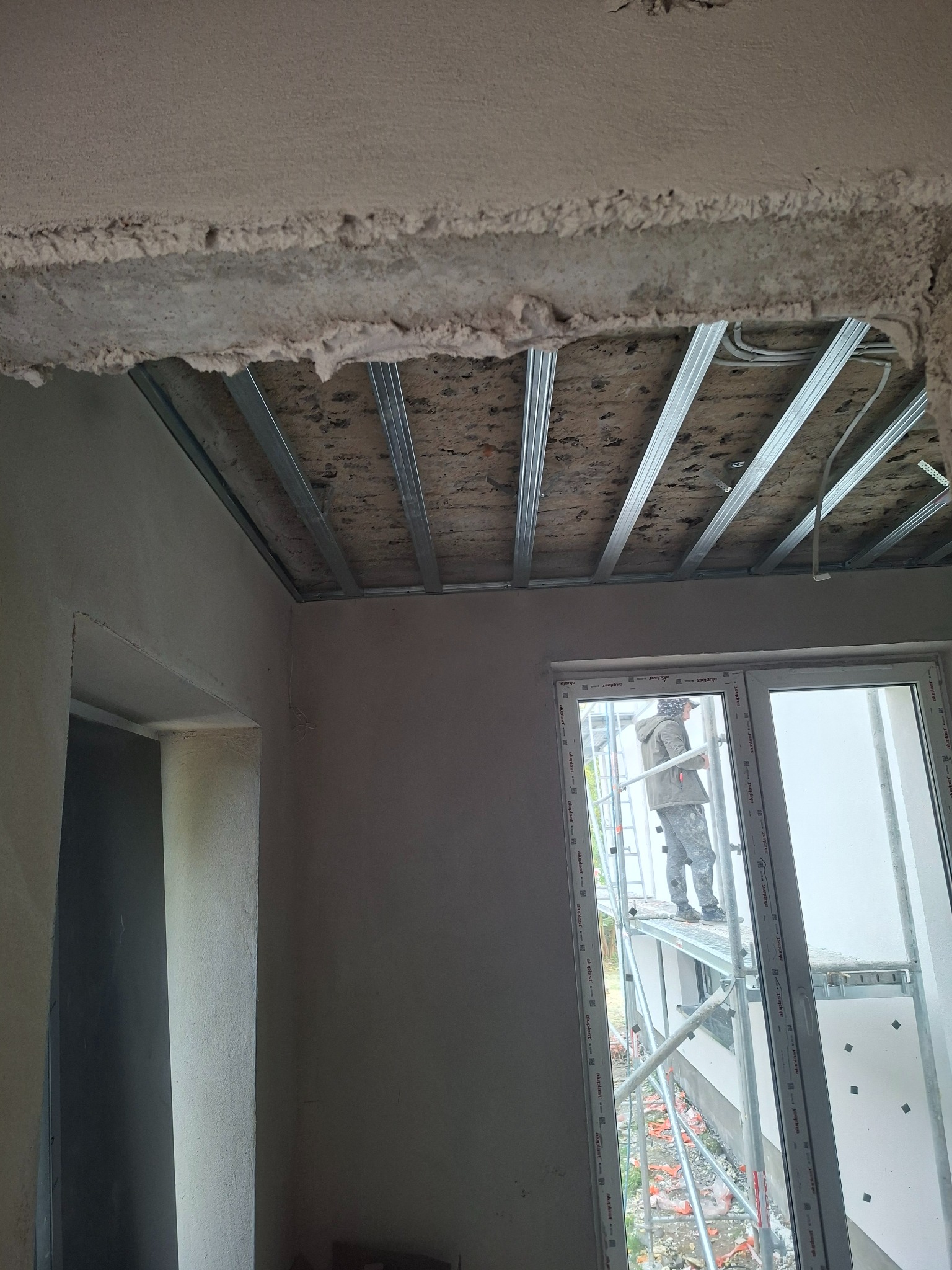 Montaż sufitu podwieszanego z metalową konstrukcją. Widoczny pracownik na rusztowaniu przy oknie. Surowy stan wykończenia wnętrza, widoczne kable i niedokończone ściany.