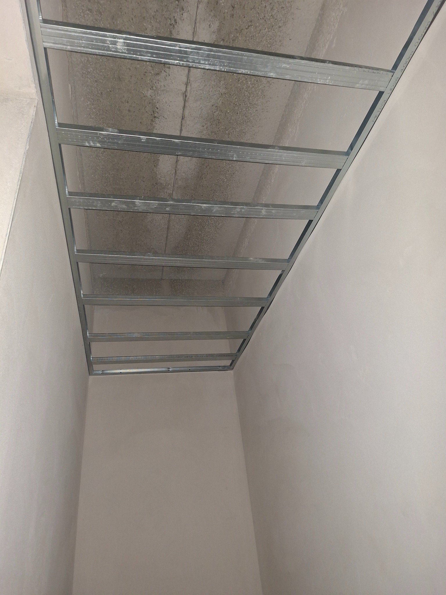 Metalowa konstrukcja sufitu podwieszanego w Śmigiel, widok z dołu. Szara ściana i surowy betonowy strop w tle. Przygotowanie do montażu płyt.