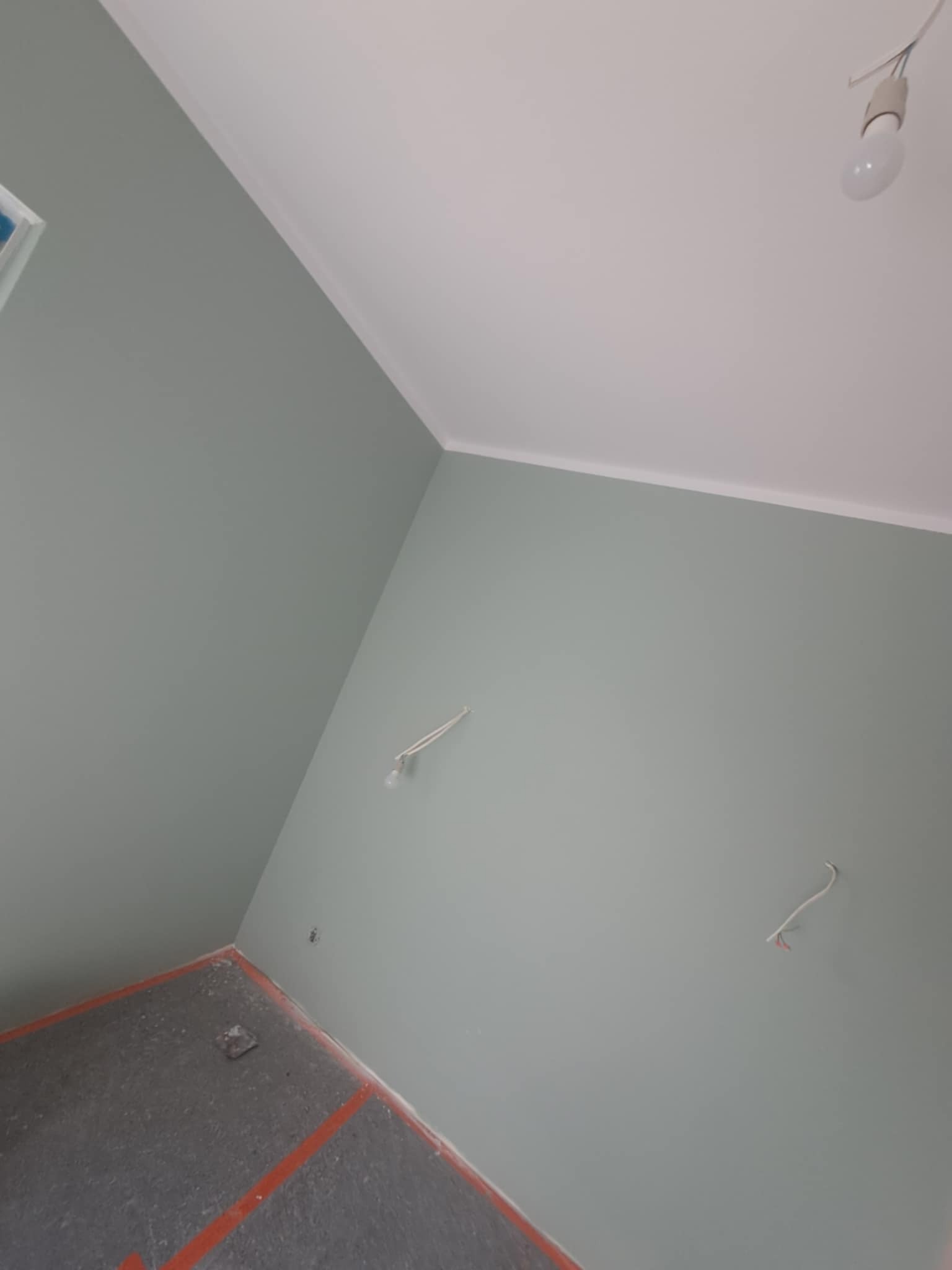 Świeżo pomalowane ściany w odcieniach szarości i bieli, widoczne wystające kable elektryczne i fragment podłogi z pomarańczową taśmą. Ujęcie z dołu.