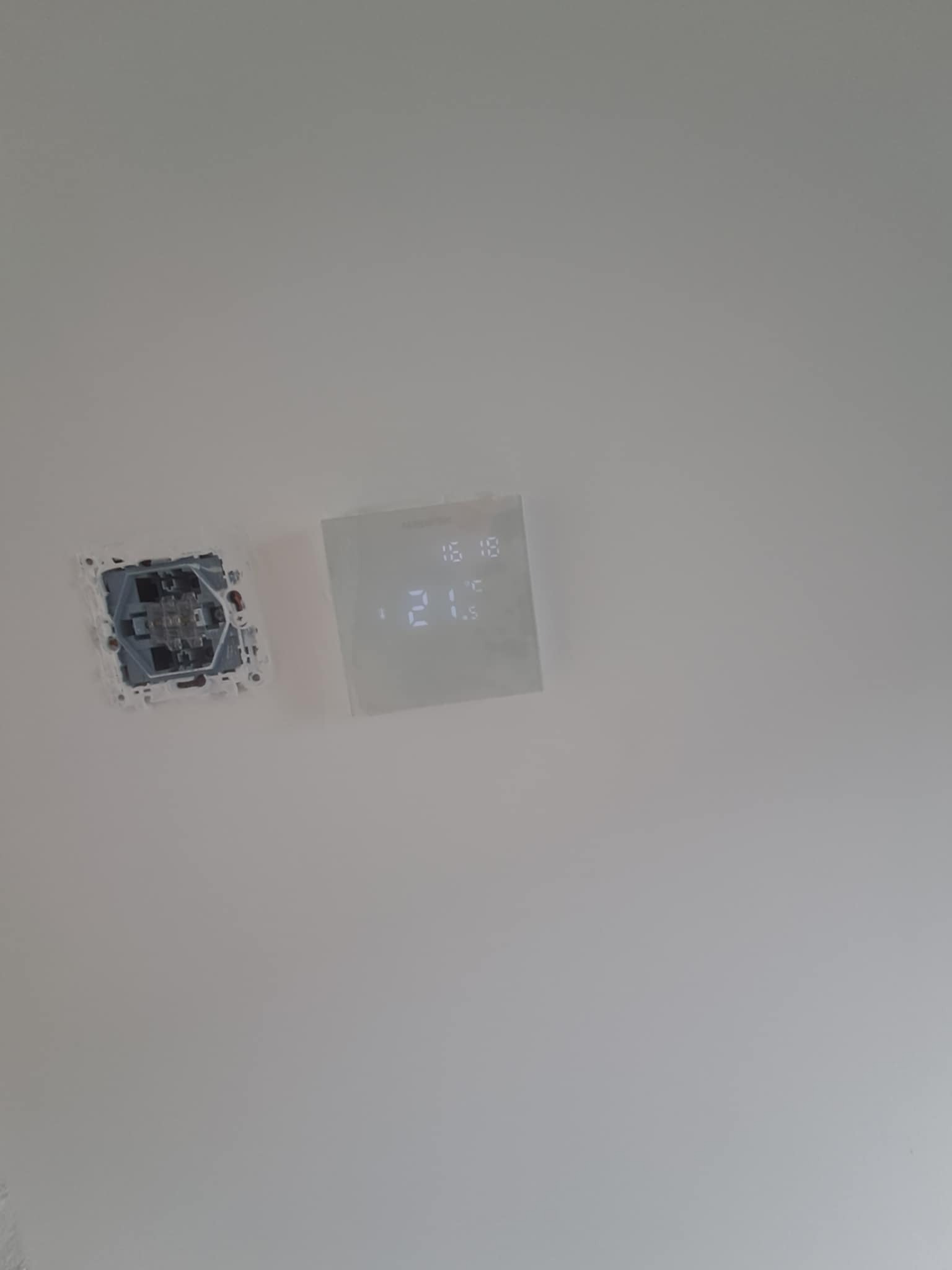 Biała ściana z zamontowanym, nowoczesnym termostatem cyfrowym i puszką elektryczną bez osprzętu. Widoczne przewody.