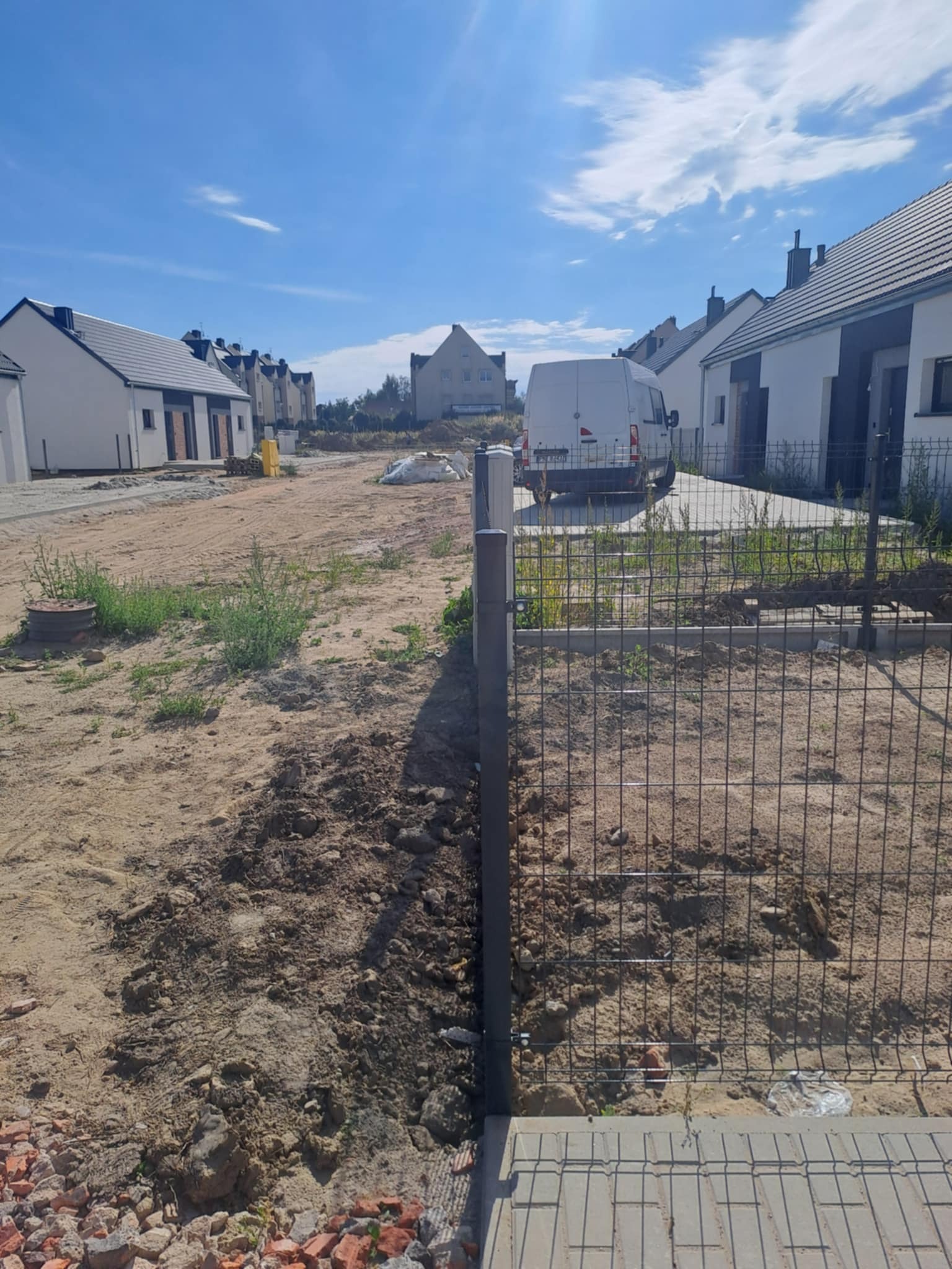 Montaż ogrodzenia panelowego na nowym osiedlu domów jednorodzinnych. Widoczne słupki i siatka ogrodzeniowa, przygotowany teren pod dalsze prace. Słoneczny dzień.