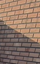 Częściowy widok ściany z czerwonej cegły, oświetlonej z góry, z wyraźnym cieniem ukośnym, podkreślającym fakturę cegieł i spoin.