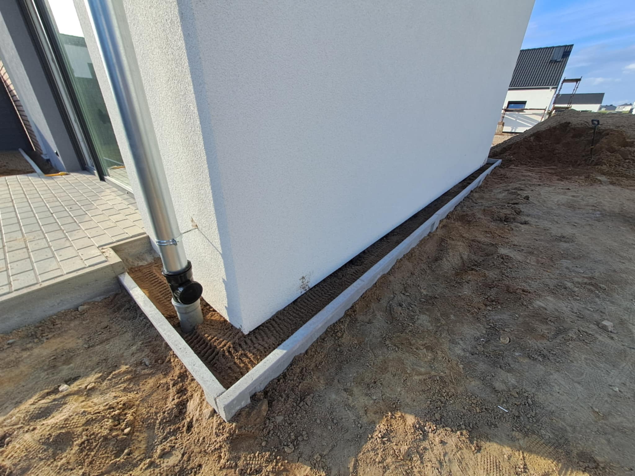 Świeżo wykończona elewacja domu z zamontowanym systemem odprowadzania wody deszczowej i obrzeżami, widok z poziomu gruntu, w tle fragment budynku.