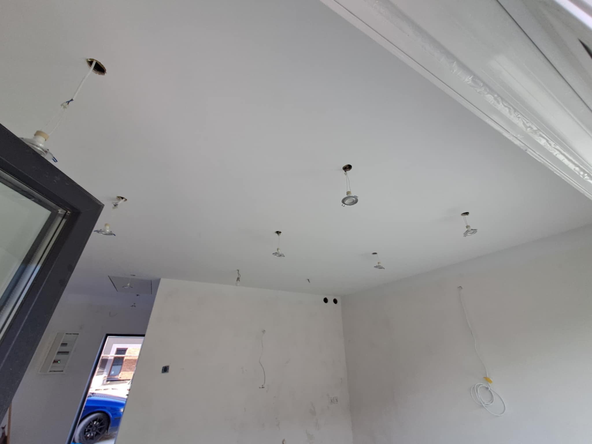 Białe wnętrze w trakcie prac wykończeniowych: sufit z otworami na oświetlenie, ściany przygotowane do malowania, widoczne przewody elektryczne.