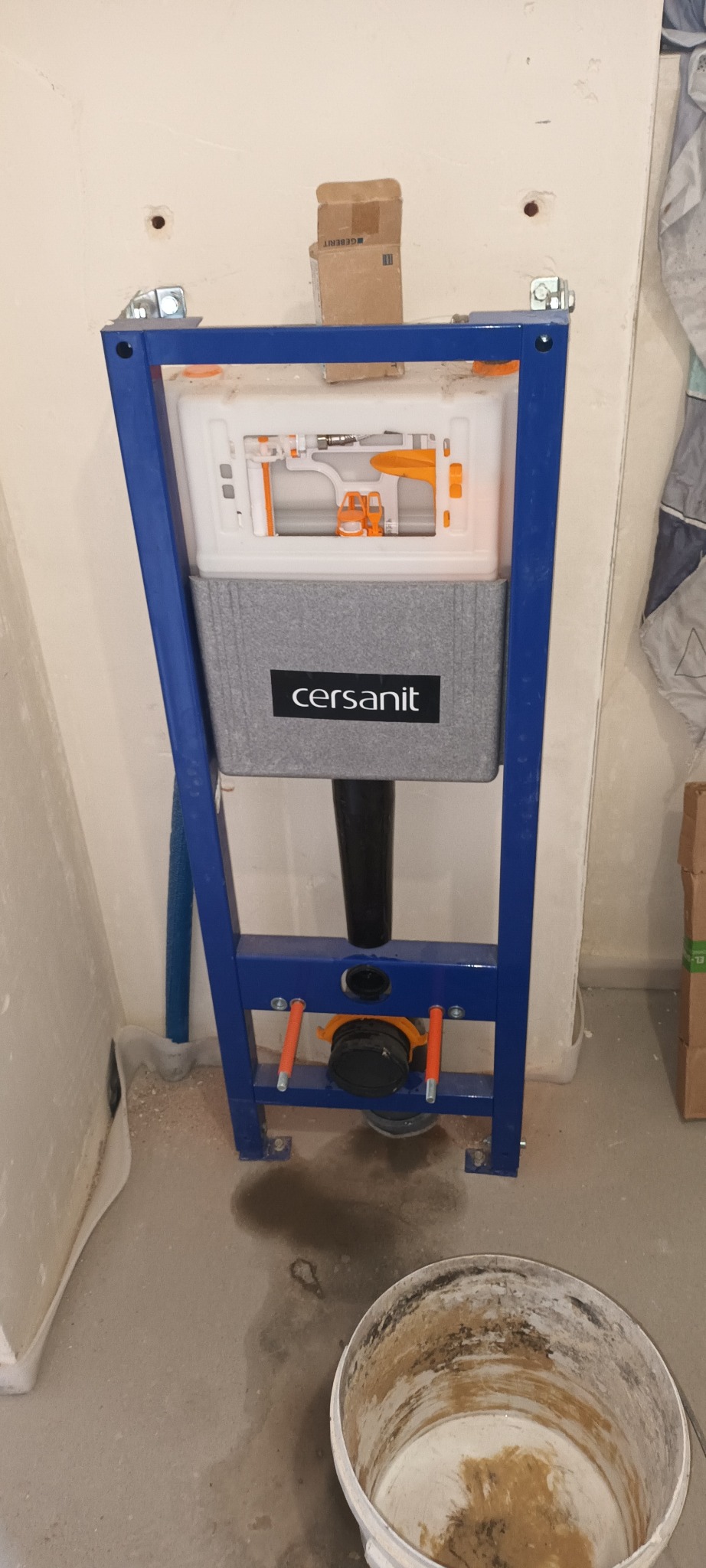 Stelaż podtynkowy Cersanit zamontowany przy ścianie, widoczny fragment instalacji wodnej i wiadro z zaprawą na podłodze. Etap prac instalacyjnych w łazience.