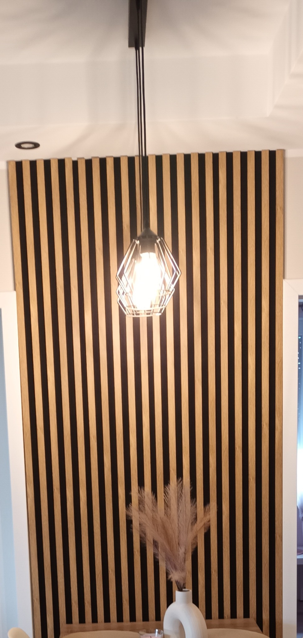 Panel ścienny z pionowych listew w kolorze drewna i czerni, oświetlony nowoczesną lampą wiszącą. Dekoracja wnętrza z wazonem i suszem na pierwszym planie.