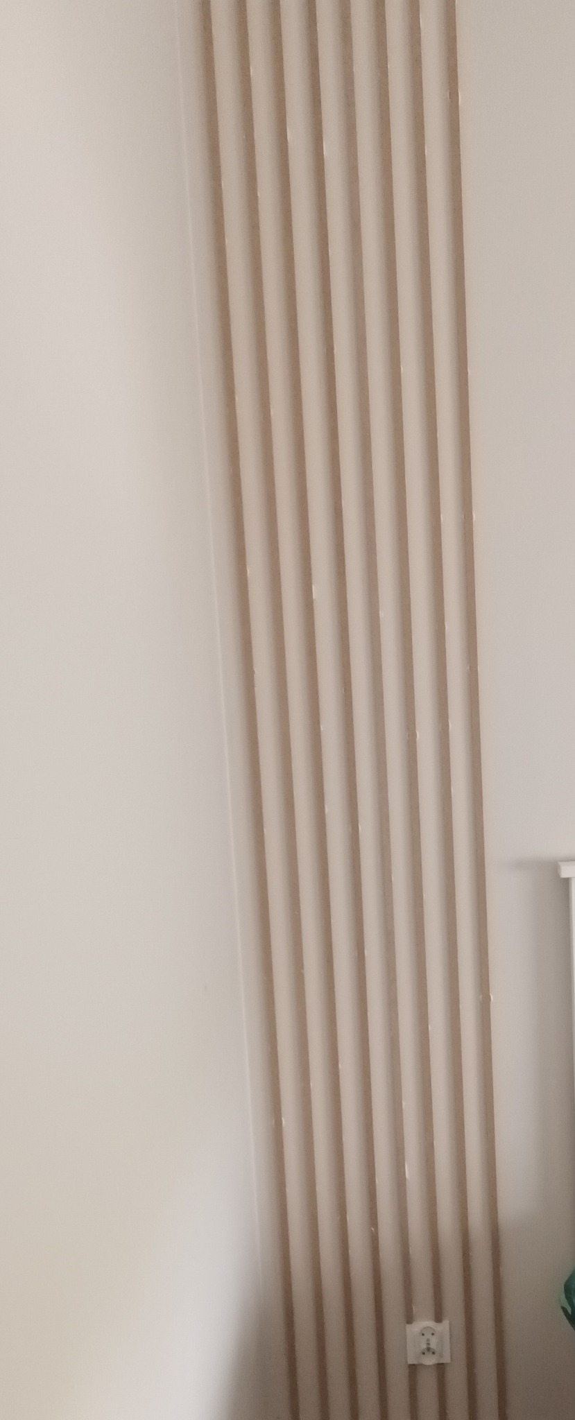 Ściana z dekoracyjnymi, pionowymi lamelami w odcieniach beżu, tworzącymi nowoczesny akcent w jasnym wnętrzu. Widoczny fragment gniazdka elektrycznego.