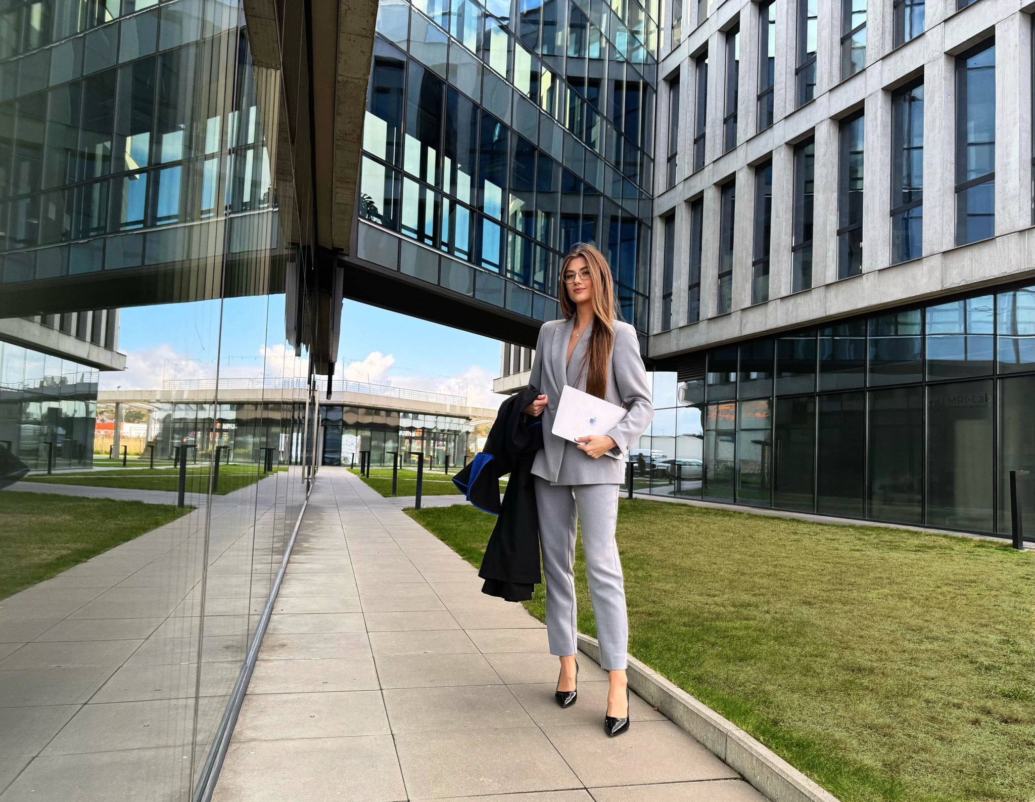 Profesjonalna prawniczka w szarym garniturze, trzymająca laptopa i togę, pozuje na tle nowoczesnego biurowca z zielonym trawnikiem. Elegancki wizerunek.