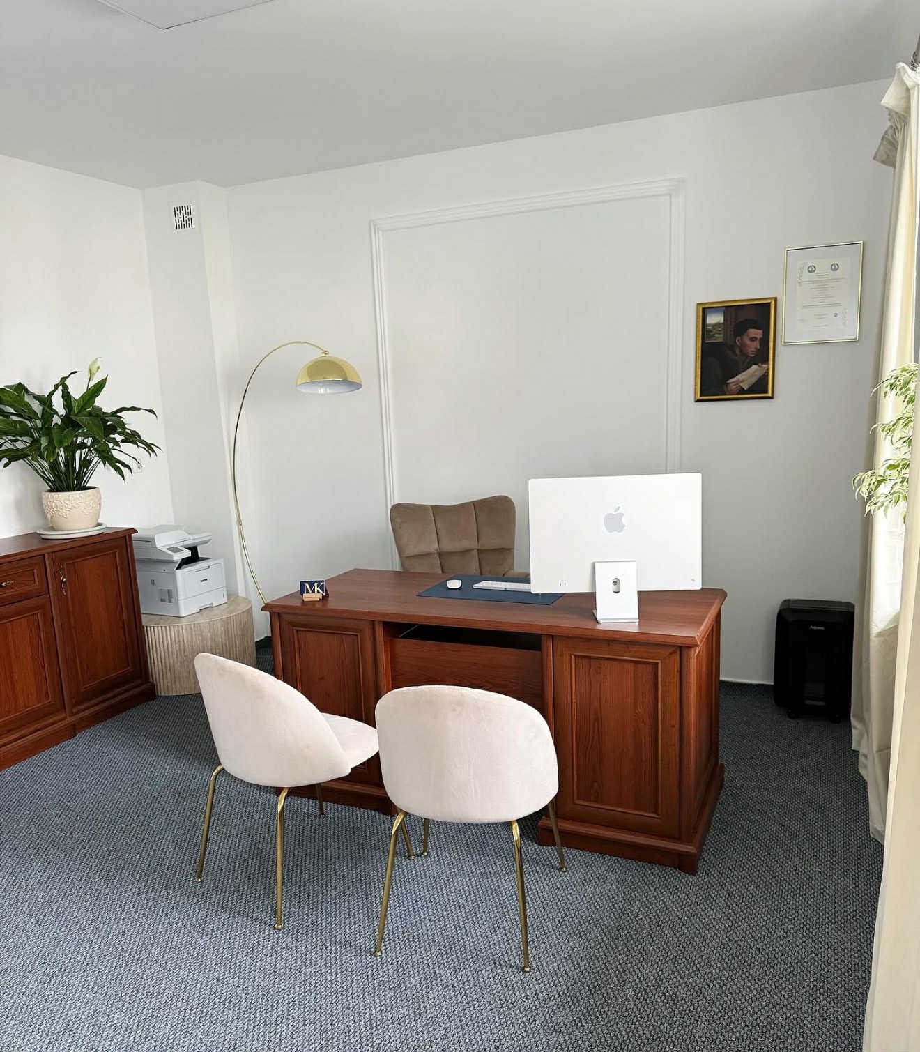 Eleganckie biuro z drewnianym biurkiem, krzesłami na złotych nogach i komputerem Apple. Na ścianie obraz i certyfikat. W tle roślina doniczkowa i lampa łukowa.