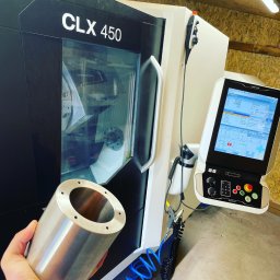 KD Technology Kacper Dulewicz - Metalowy cylinder z otworami trzymany na tle nowoczesnej maszyny CLX 450. Widoczny panel sterowania i wnętrze maszyny z obrabianym elementem.
