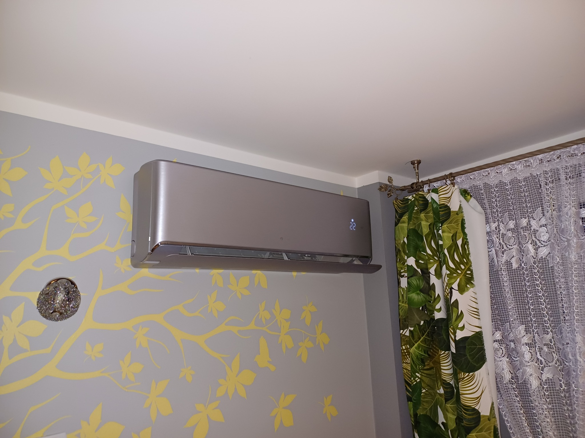 Zamontowany srebrny klimatyzator na ścianie z motywem liści, obok okno z zasłonami w tropikalne wzory i koronkową firanką. Widoczny fragment sufitu i lampy.
