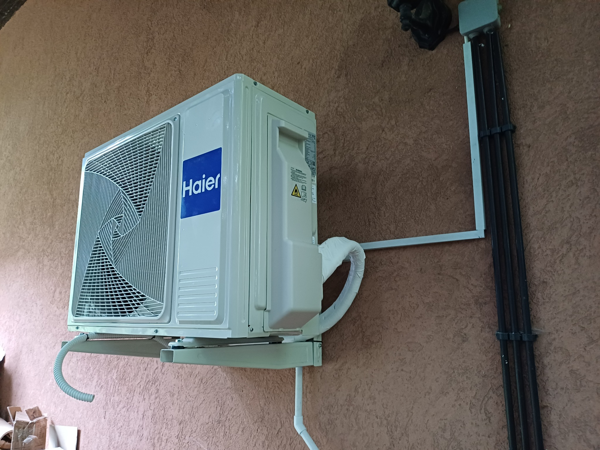 Zamontowany na zewnątrz klimatyzator Haier na brązowej ścianie. Widoczne rury odprowadzające i przewody elektryczne w listwie. Ujęcie z dołu.