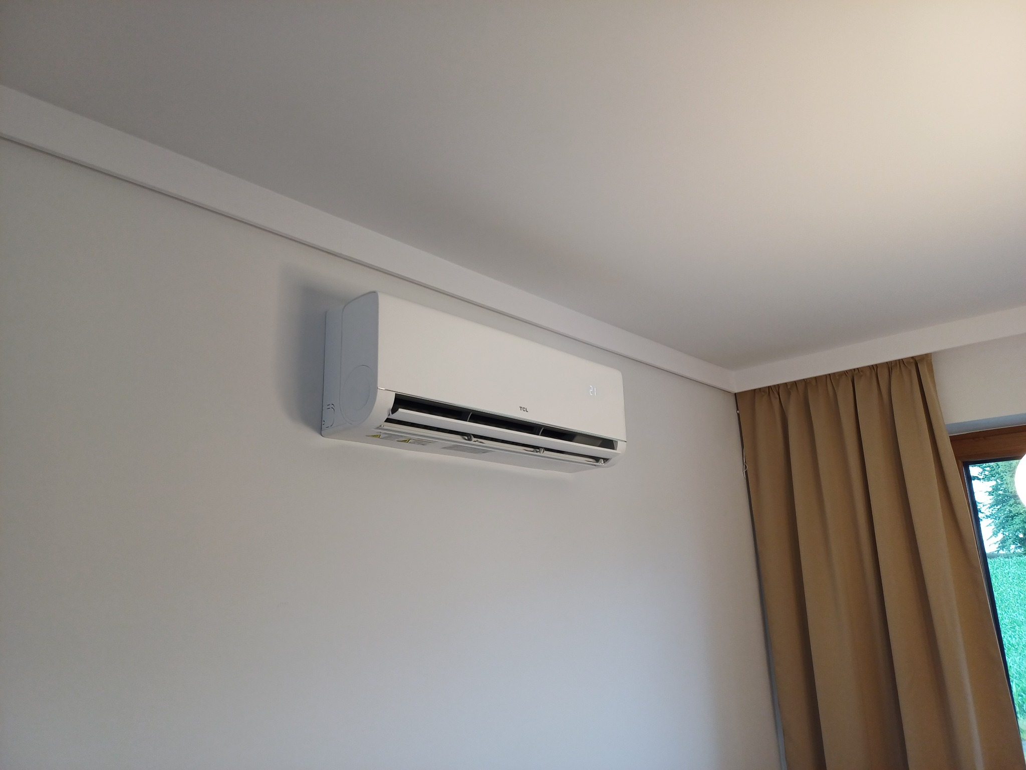 Biały klimatyzator TCL zamontowany na ścianie w jasnym pokoju, widoczne brązowe zasłony i fragment okna. Minimalistyczny design, nowoczesne wnętrze.