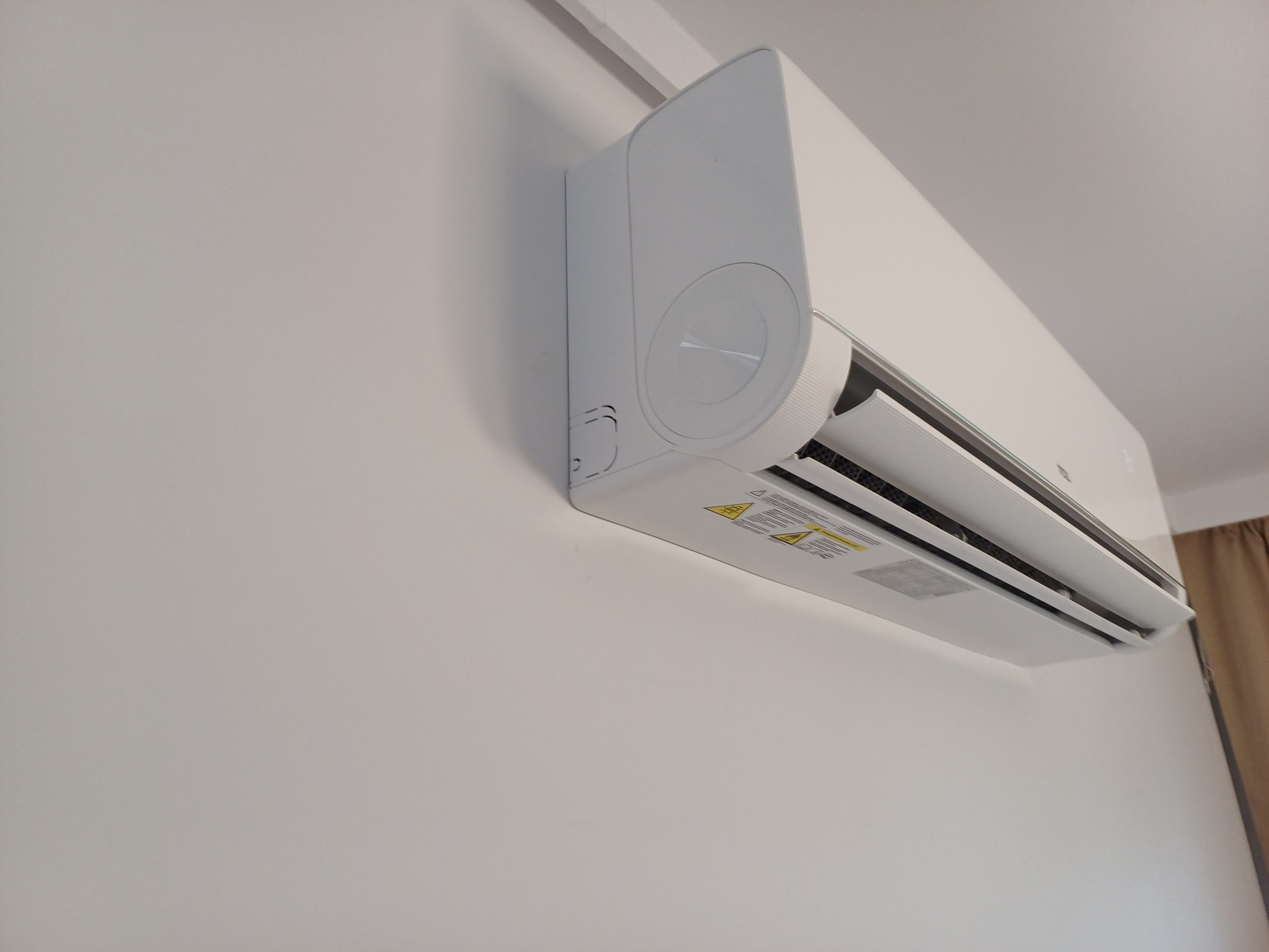 Biały klimatyzator ścienny zamontowany w pomieszczeniu, widoczny z boku, z fragmentem zasłony w tle. Minimalistyczny design, czyste linie, nowoczesny wygląd.