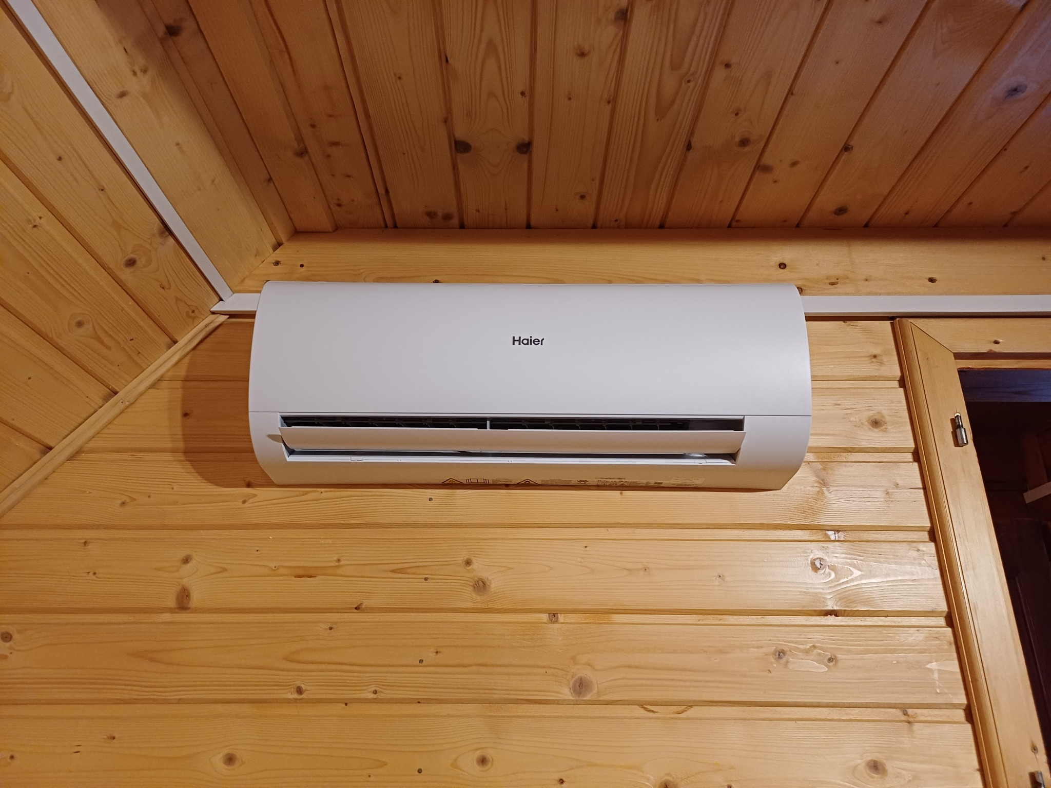 Biały klimatyzator Haier zamontowany na ścianie z jasnego drewna wewnątrz pomieszczenia. Widoczny fragment drewnianego sufitu i futryny drzwi.