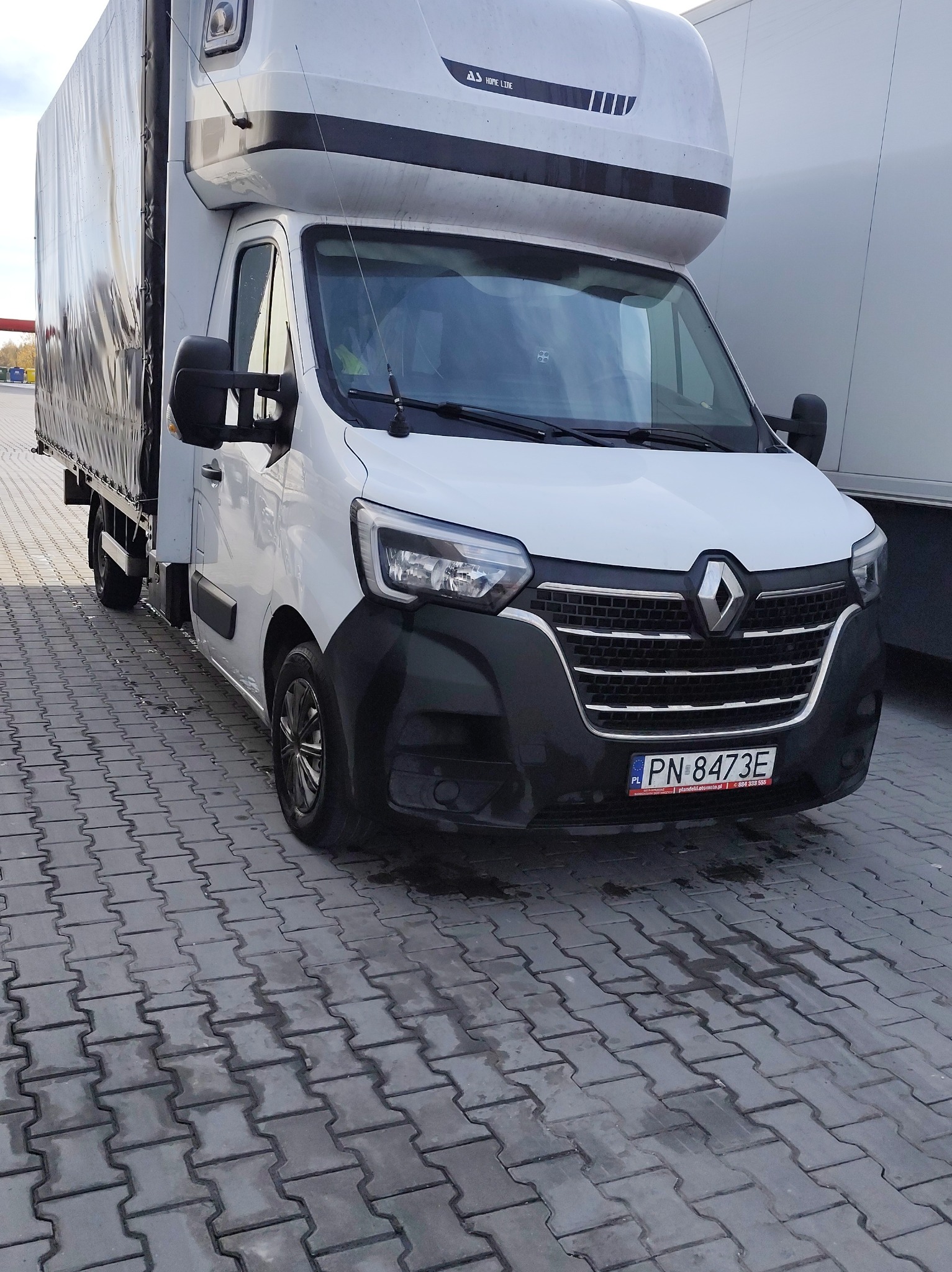Biały bus Renault Master z plandeką na parkingu z kostki brukowej. Widoczna tablica rejestracyjna. Auto gotowe do transportu.