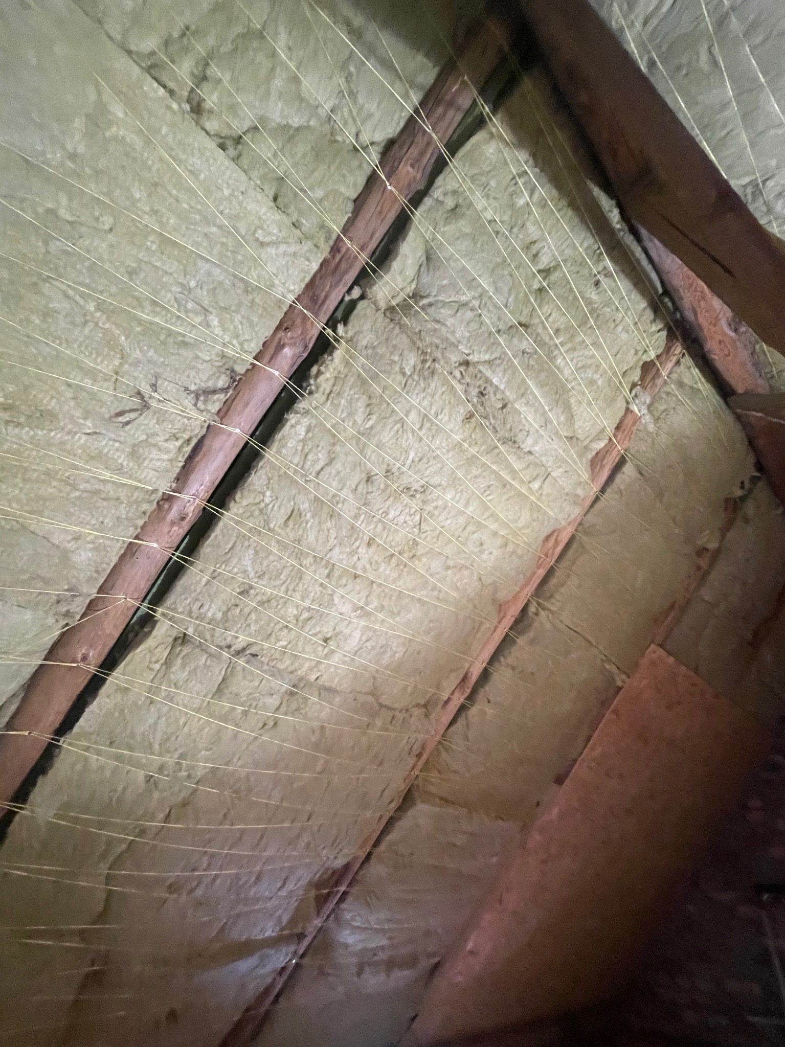 Poddasze z widoczną izolacją z wełny, zabezpieczoną sznurkami. Elementy drewniane konstrukcji dachu. Ujęcie z dołu, podkreślające strukturę materiału izolacyjnego.