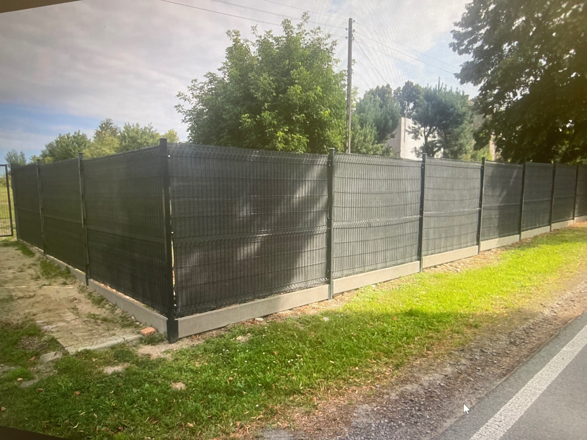 Nowoczesne ogrodzenie panelowe w kolorze grafitowym, z podmurówką betonową, otoczone zielenią. Widok z zewnątrz posesji, w tle drzewa i błękitne niebo.
