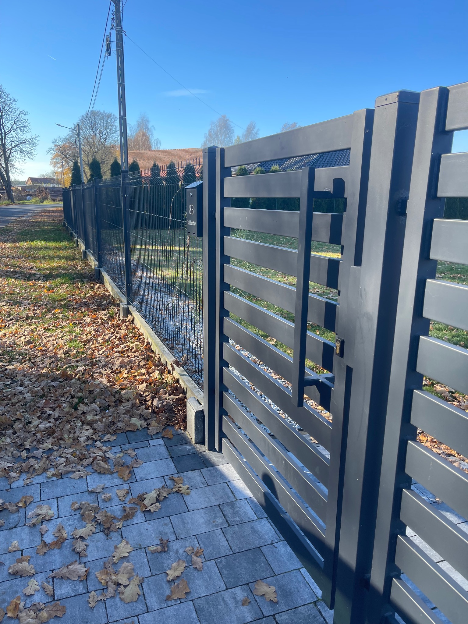 Nowoczesne, grafitowe ogrodzenie panelowe z furtką, widok z zewnątrz na posesję. Na pierwszym planie kostka brukowa pokryta opadłymi liśćmi.
