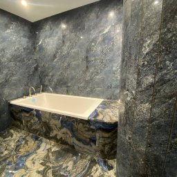 Kosmina Group - Wykończona łazienka z wanną obudowaną płytkami imitującymi marmur w odcieniach szarości i błękitu. Płytki na ścianach i podłodze tworzą spójną, elegancką całość.