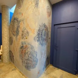 Ściana z dekoracyjnym malowaniem w odcieniach błękitu i beżu, wzory mandali, obok niebieskie drzwi i kolumna z mozaiki. Wykończenie wnętrza z dbałością o detale.