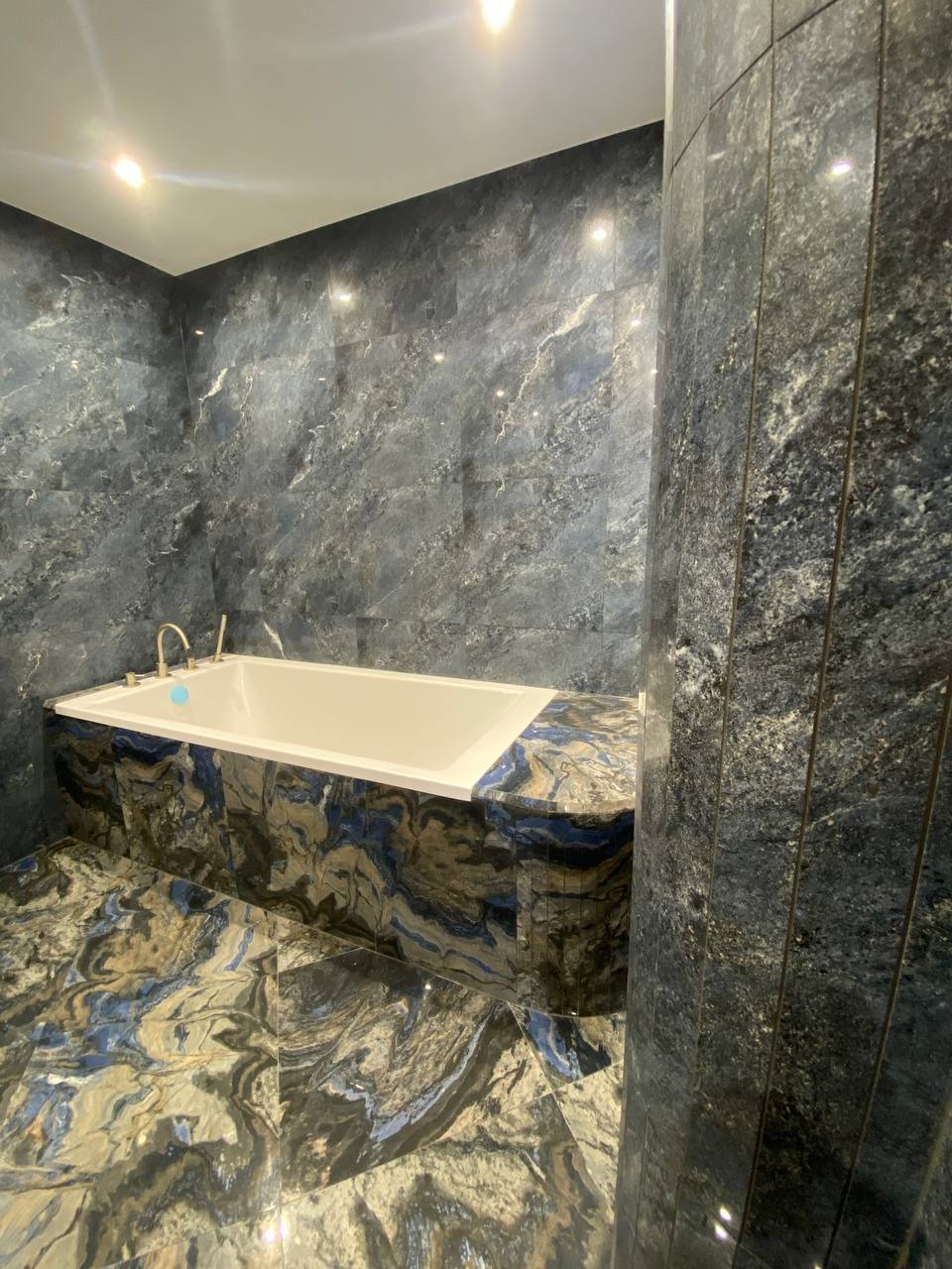 Wykończona łazienka z wanną obudowaną płytkami imitującymi marmur w odcieniach szarości i błękitu. Płytki na ścianach i podłodze tworzą spójną, elegancką całość.