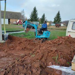 Piotr Dąbrowa - Niebieska mini koparka Kingway z operatorem w trakcie prac ziemnych na działce. W tle budynek gospodarczy i drzewa iglaste.