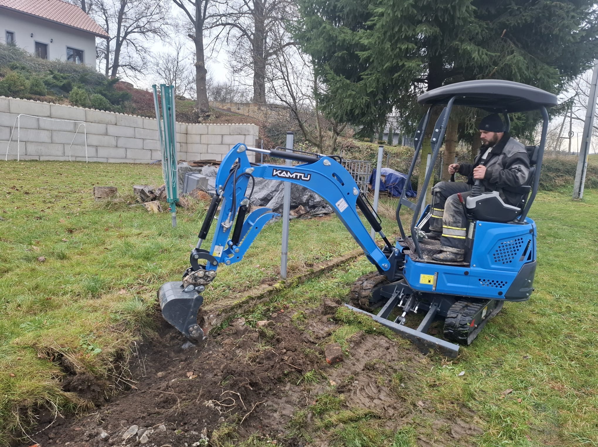 Niebieska mini koparka KAMTU w trakcie wykonywania wykopu w ziemi na trawiastym terenie. Operator w kabinie. W tle mur z betonowych bloków i drzewa.