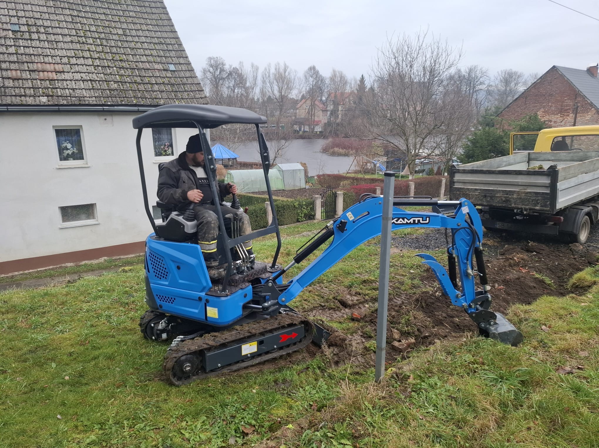 Niebieska mini koparka KAMTU w akcji, operator w kabinie, wykop w trawie, obok zaparkowana ciężarówka, w tle zabudowa wiejska i zbiornik wodny.