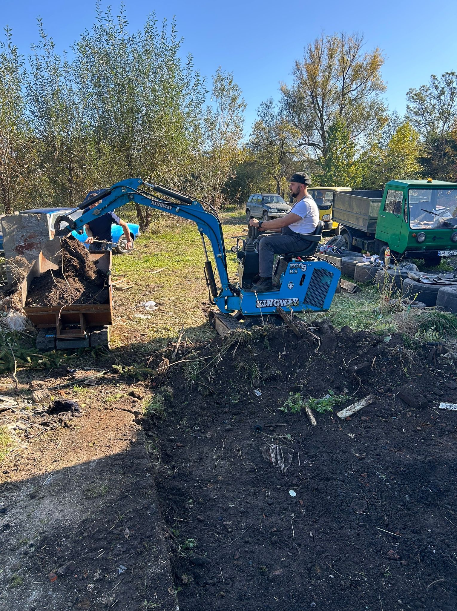 Niebieska mini koparka Kingway w trakcie pracy, operator w białej koszulce, wykopana ziemia w łyżce, w tle zieleń i pojazdy. Prace ziemne w słoneczny dzień.